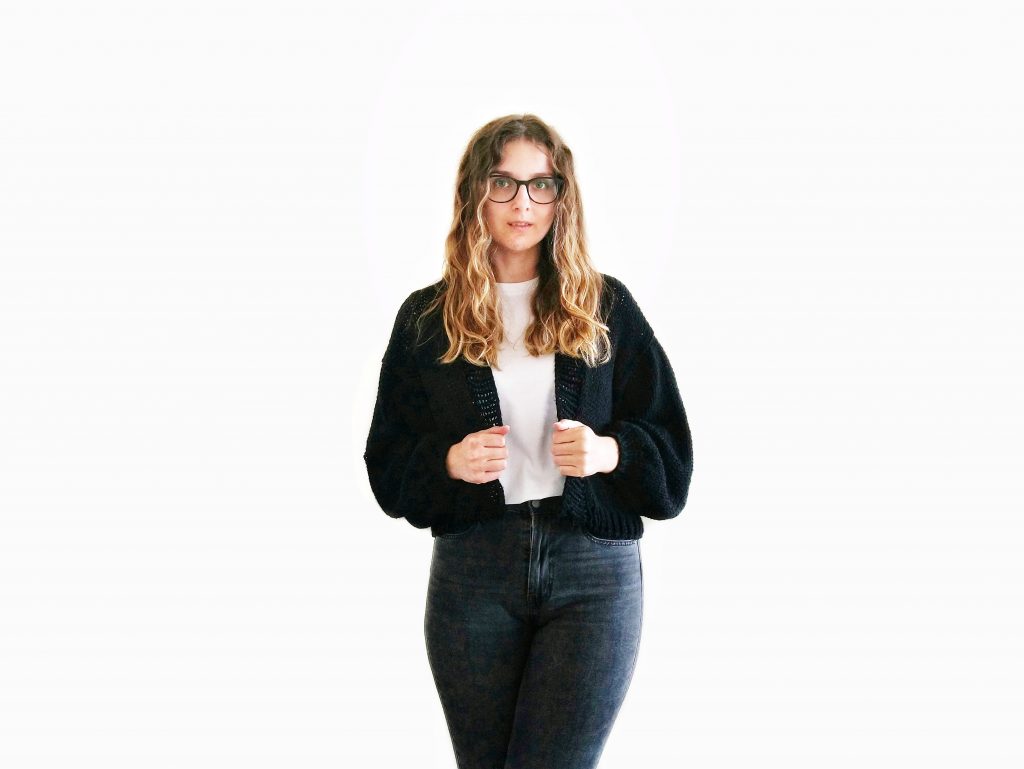 A photo of Jess against a white background wearing jeans, a white t-shirt and a black, cropped knit cardigan with loose sleeves, hand knit from Knitatude's Top Gun Bomber pattern