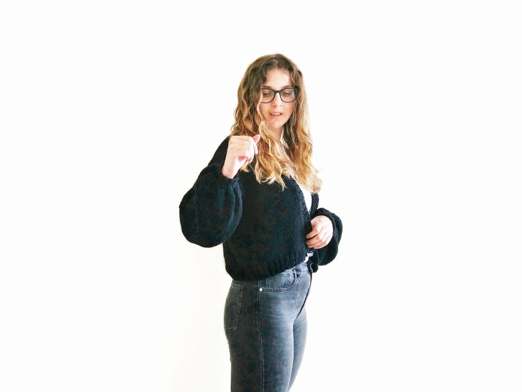 A photo of Jess against a white background wearing jeans, a white t-shirt and a black, cropped knit cardigan with loose sleeves, hand knit from Knitatude's Top Gun Bomber pattern